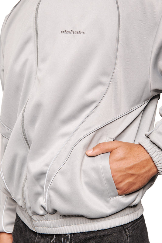 Person trägt die Rave Trackjacket Gray mit Stehkragen, Reißverschluss, reflektierenden Teilungsnähten, elastischen Bündchen und gesticktem olahala Logo auf der Brust.