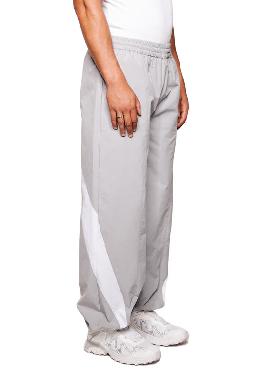 Person trägt OLAKALA Sports Trackpants Gray mit weißen Streifen, elastischem Bund und gesticktem Logo auf linkem Bein, kombiniert mit weißen Sneakers.
