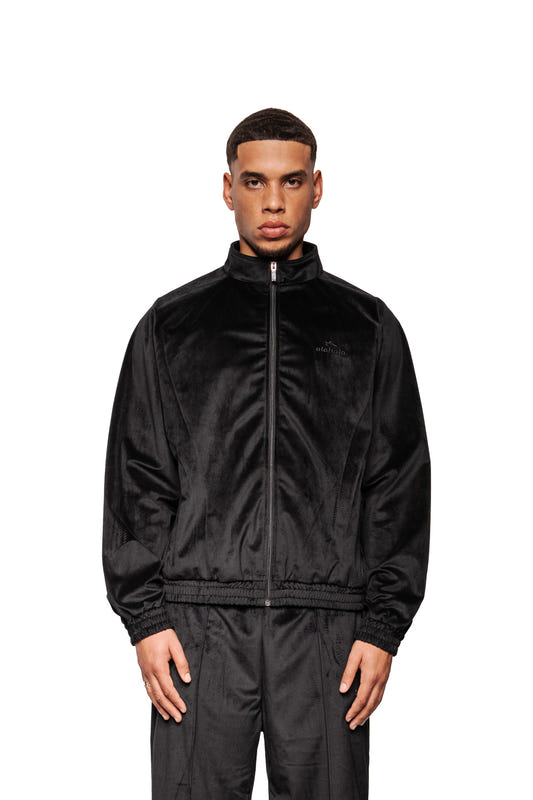 Männliches Modell in Sports Velour Jacket Black mit hohem Kragen, Raglanärmeln, elastischem Saum; lockere, nahtbetonte Hose; dezentes Logo links auf der Brust.