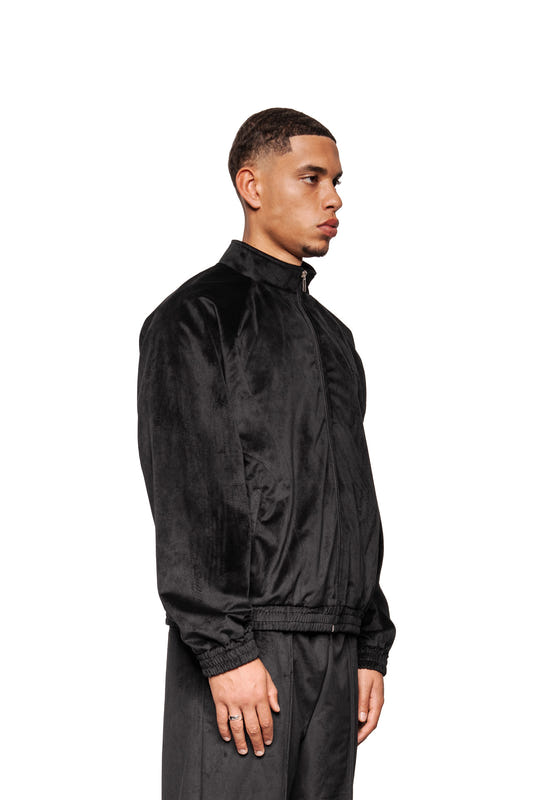 Männliches Modell im Profil trägt eine schwarze Sports Velour Jacke mit sportlichen Teilungsnähten, weichem Velours und atmungsaktivem Mesh-Futter, präsentiert von OLAKALA.