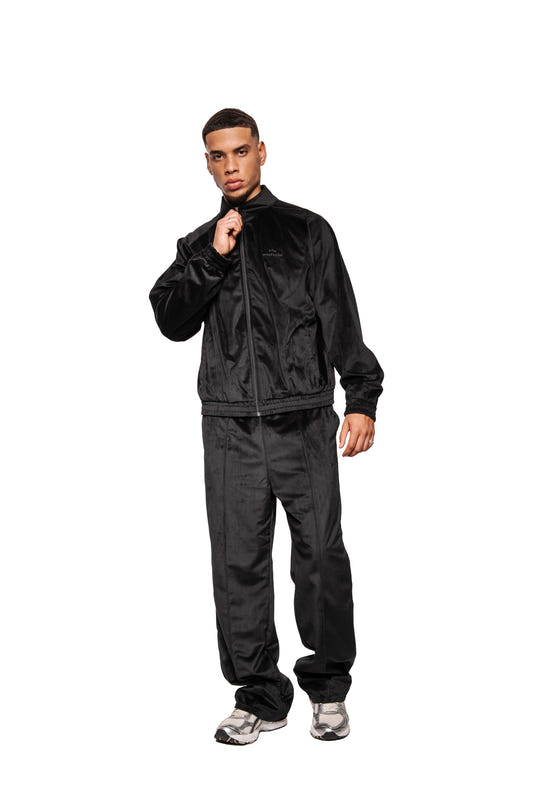 Männliches Modell in Sports Velour Jacket Black aus hochwertigem Velours mit Teilungsnähten, elastischen Bündchen und lockerem Schnitt, kombiniert mit passenden Trackpants und Sneakern.