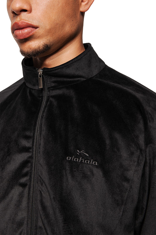 Person in schwarzer Sports Velour Jacket Black mit Stehkragen, Reißverschluss und kleinem otahala-Logo, zeigt sportlich-modernen Look durch Teilungsnähte, weiches Velours und lockere Passform.