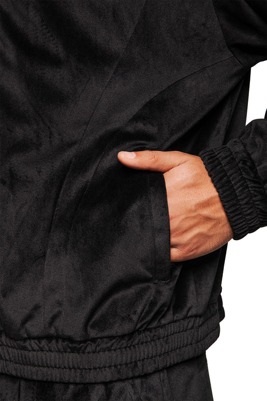 Person trägt die Sports Velour Jacket Black mit elastischen Bündchen und Seitentaschen, aus weichem Velours-Material, das sportlich-modernen Look und Komfort bietet.