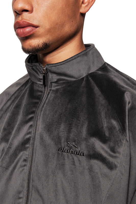 Nahaufnahme einer Person, die eine grau-olakala Sports Velour Jacke mit Stehkragen und gesticktem Logo trägt. Velours-Material und Mesh-Futter sorgen für Komfort und sportlichen Look.