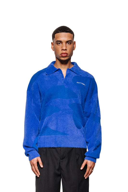 Mann trägt Synergy Glitched Fuzzy Polo Blue: langärmliger, strukturierter Pullover mit Polo-Kragen, gesticktem Logo, gerippten Bündchen, weicher Oberfläche, kombiniert mit schwarzen Hosen.