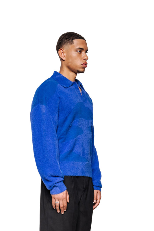 Junger Mann im Profil trägt Synergy Glitched Fuzzy Polo Blue mit Polokragen, Ripp-Bündchen, gesticktem Logo und abstraktem Muster, kombiniert mit schwarzen Hosen.