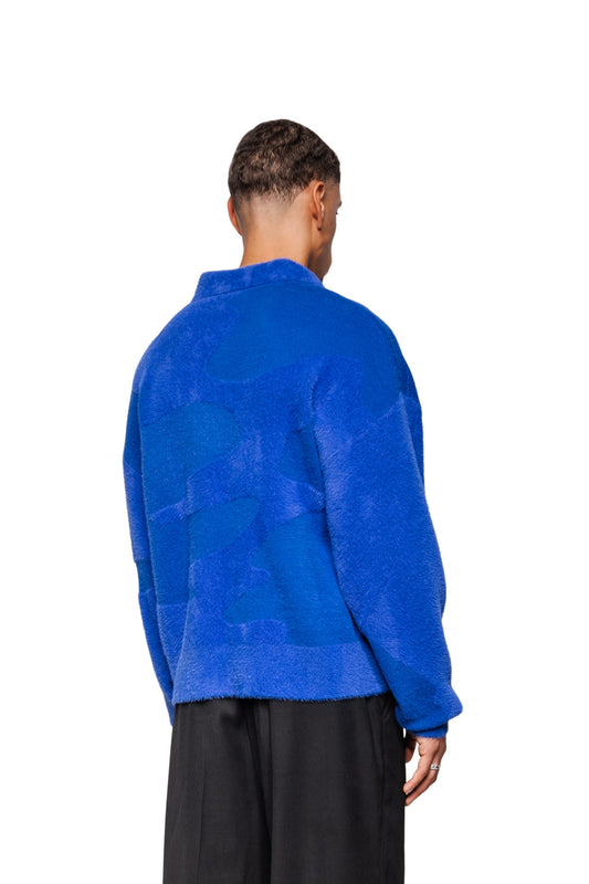 Person von hinten in Synergy Glitched Fuzzy Polo Blue mit Polo-Kragen, Ripp-Bündchen und gesticktem Logo, getragen mit lockerer schwarzer Hose.