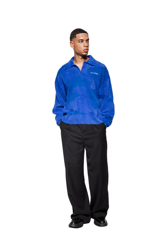 Junger Mann trägt Synergy Glitched Fuzzy Polo Blue mit abstraktem Muster, Polo-Kragen, Ripp-Bündchen, gesticktem Logo auf der Brust, kombiniert mit schwarzen Hosen.