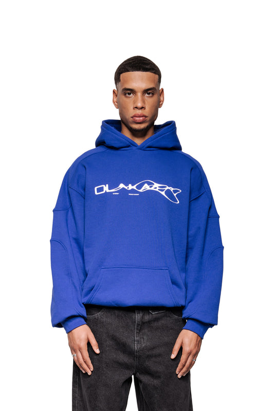 Junger Mann trägt Synergy Mask Hoodie Blue mit Kängurutasche und abstraktem Logo. Oversized und unisex, kombiniert mit faded Jeans, betont durch Ripp-Bündchen und Teilungsnähte.