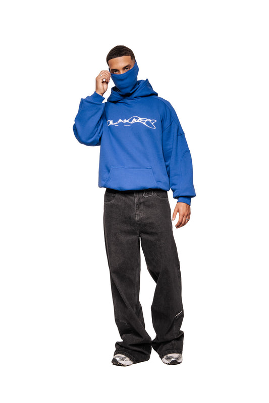 A young man wears the Synergy Mask Hoodie Blue with integrated face protection, white chest print and kangaroo pocket, combined with loose-fitting jeans and sneakers.