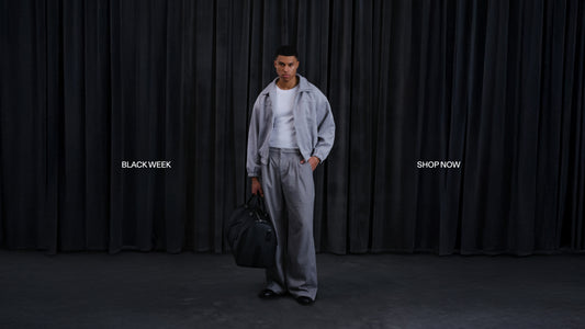 Mann in grauem Freizeit-Outfit mit Reisetasche vor schwarzem Vorhang; Banner „Black Week“ im Hintergrund.