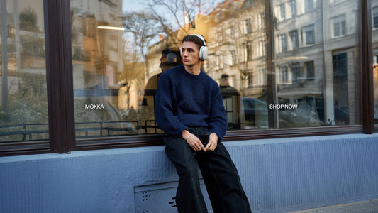 Mann in dunkelblauem Strickpullover und schwarzen Hosen sitzt vor dem Schaufenster eines Stadtladens, trägt Kopfhörer; urbane Straßenszene.