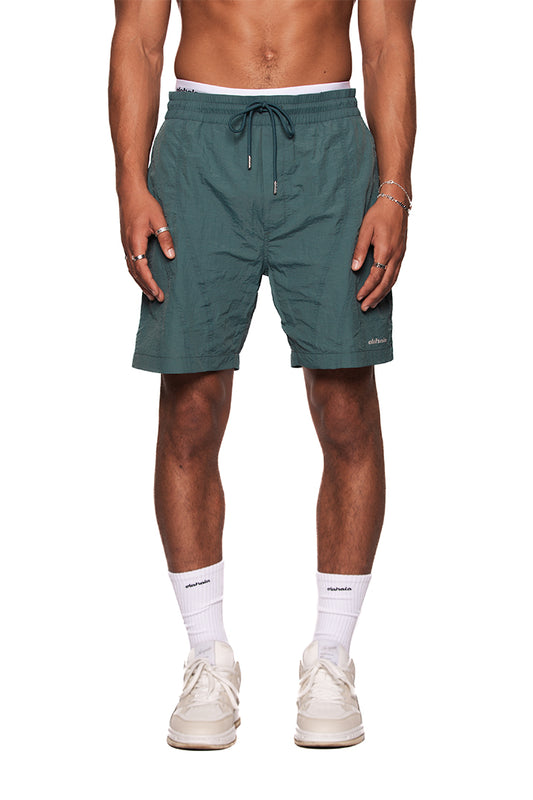 Mann trägt Naxos Swim Shorts mit verstellbarem Bund und reflektierendem OLAKALA-Logo, kombiniert mit weißen Sneakers und Socken.