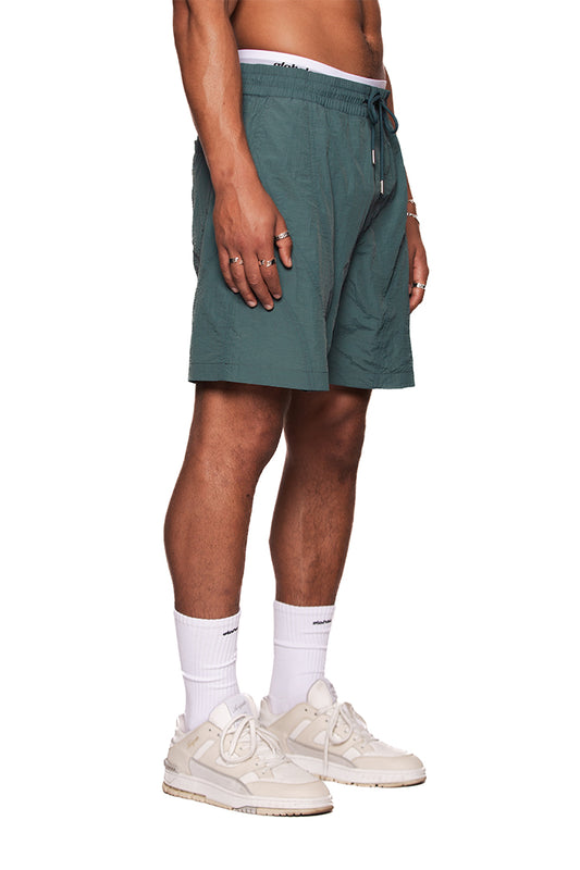 Person trägt Naxos Swim Shorts mit Teilungsnähten, verstellbarem Bund und reflektierendem OLAKALA-Logo, kombiniert mit weißen Socken und Sneakers.