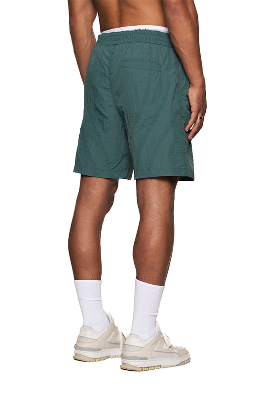 Person trägt Naxos Swim Shorts Green mit verstellbarem Bund und reflektierendem OLAKALA-Logo, zeigt Bein- und Fußbereiche.