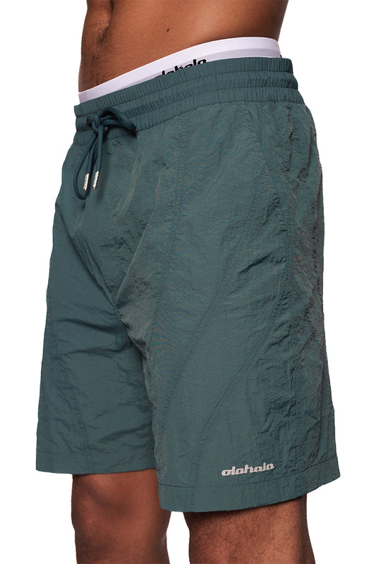Person trägt Naxos Swim Shorts Green mit Teilungsnähten, verstellbarem Bund, französischen Taschen und reflektierendem OLAKALA Logo auf dem linken Bein.