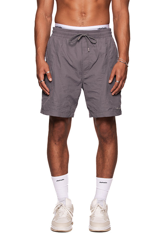 Mann trägt Naxos Swim Shorts Gray mit verstellbarem Bund und reflektierendem OLAKALA-Logo, kombiniert mit weißen Sneakers und Socken.
