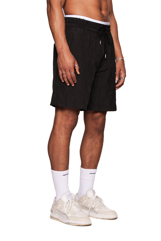 Person trägt die Naxos Swim Shorts Black mit Teilungsnähten, verstellbarem Bund und reflektierendem OLAKALA Logo, kombiniert mit weißen Sneakers und Socken.