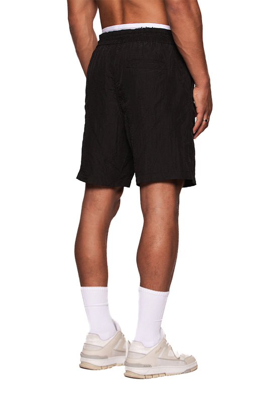 Person trägt Naxos Swim Shorts Black mit verstellbarem Bund und reflektierendem OLAKALA-Logo auf dem linken Bein, kombiniert mit weißen Socken und Sneakern.